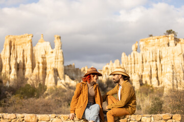 A man and woman are sitting on a stone wall in front of a mountain. The woman is wearing a cowboy...