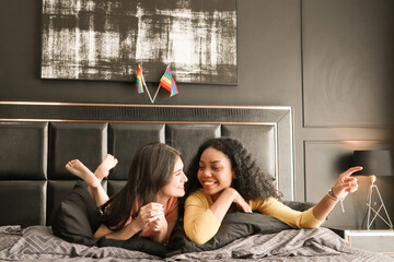 A black young woman pointing and looking out the window at home. Attractive happy teenage lesbian couples spend romantic time in the bedroom with rainbow flags behind them, a weekend getaway concept.