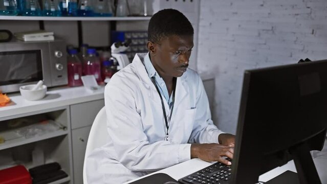 An african man in a lab coat works intently at a computer in a modern laboratory setting.