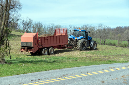 Blue tractor pulls a red fertilizer manure spreader over a green early spring agricultural field by a country road under a blue sky