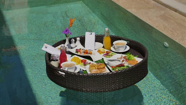 Luxury floating breakfast in exotic swimming pool at tropical resort. Sumptuous meal with fresh fruits, beverages, and flowers served on woven tray. Experiencing upscale travel and dining.