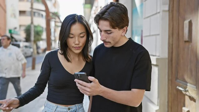 A young interracial couple engrossed in a smartphone on a bustling city street.