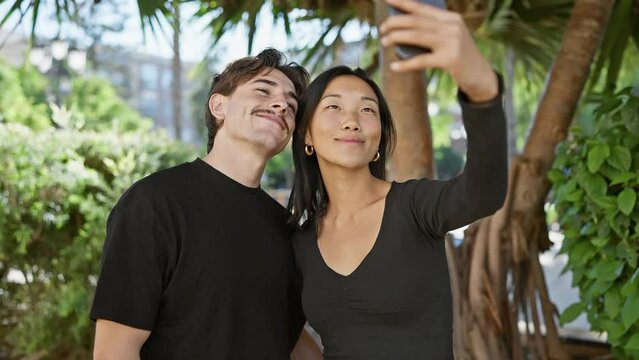 Interracial couple taking a selfie outdoors with a smartphone, showcasing their relationship and joy in a natural urban park.