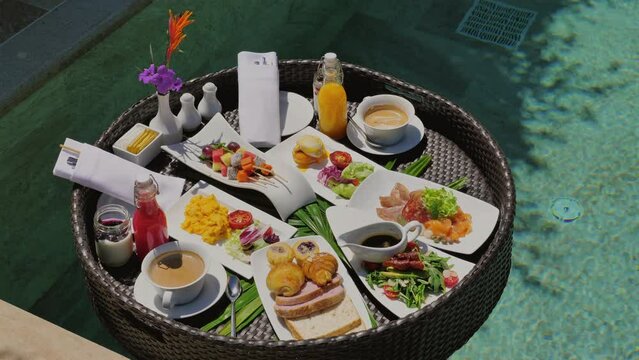 Luxurious poolside breakfast with variety of gourmet foods served on tray featuring fresh fruits, pastries, and beverages. Tropical resort dining experience.