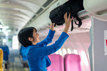 A traveler in a blue jacket is placing her carry-on bag into the overhead storage compartment on an...