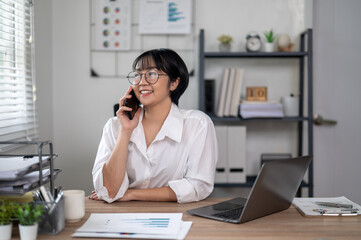 Cheerful businesswoman in white shirt using smartphone with laptop and documents on her office desk.