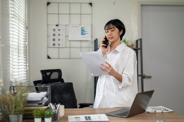 Cheerful businesswoman in white shirt using smartphone with laptop and documents on her office desk.