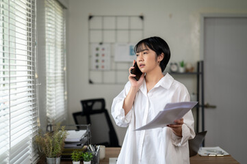 Cheerful businesswoman in white shirt using smartphone with laptop and documents on her office desk.