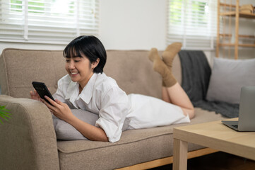 Obraz premium A smiling young woman enjoys a casual conversation on her smartphone while relaxing comfortably on a couch in a well-lit living space.