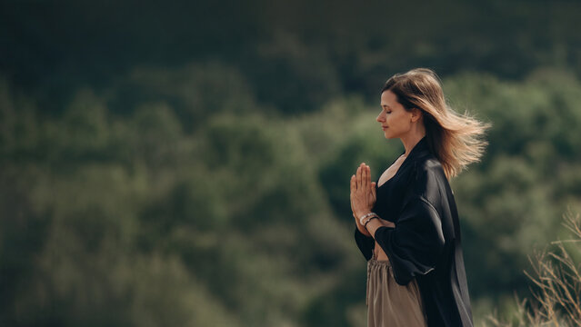 Woman and spirituality, a young female in a black kimano meditates with a namaskar gesture and contemplates nature. Side view of Caucasian woman and relaxation