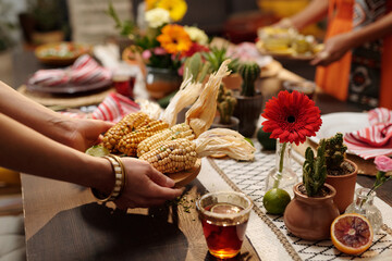 Hands of young unrecognizable Hispanic woman putting tray with homemade corn on table served for guests invited for home party - 782922925