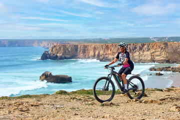 happy active senior woman cycling at the the rock cliffs and lighthouse of Cabo Sao Vicente, the south-western spit of Europe at the atlantic coast of  Algarve, Portugal,  - 782924315