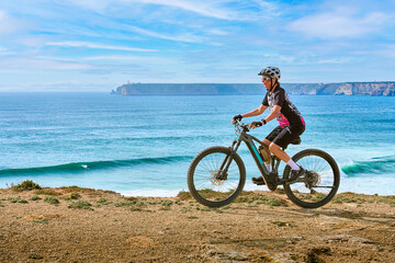 happy active senior woman cycling at the the rock cliffs and lighthouse of Cabo Sao Vicente, the south-western spit of Europe at the atlantic coast of  Algarve, Portugal,  - 782924583