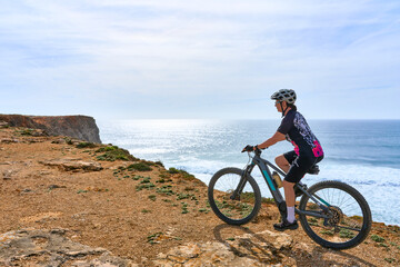happy active senior woman cycling at the the rock cliffs and lighthouse of Cabo Sao Vicente, the south-western spit of Europe at the atlantic coast of  Algarve, Portugal,  - 782924778