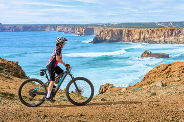 happy active senior woman cycling at the the rock cliffs and lighthouse of Cabo Sao Vicente, the south-western spit of Europe at the atlantic coast of  Algarve, Portugal,  - 782924972