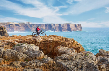 happy active senior woman cycling at the the rock cliffs and lighthouse of Cabo Sao Vicente, the south-western spit of Europe at the atlantic coast of  Algarve, Portugal,  - 782925334