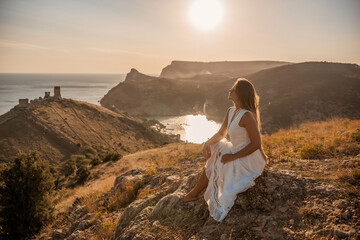 A woman in a white dress sits on a rock overlooking a body of water. The scene is serene and...