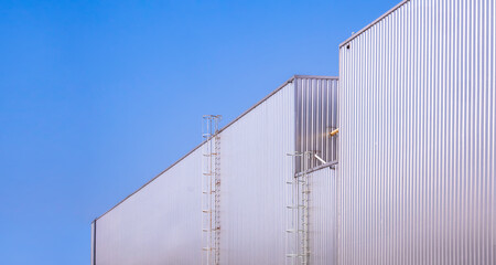 Two metal industrial warehouse buildings with cylinder ladders on aluminium corrugated wall against blue clear sky Background, perspective side view with copy space 