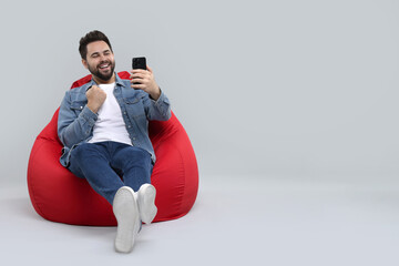 Happy young man using smartphone on bean bag chair against grey background, space for text