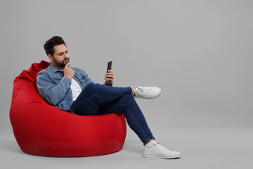 Handsome young man using smartphone on bean bag chair against grey background, space for text