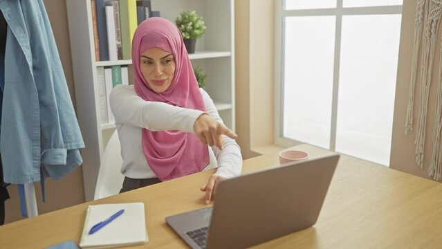 Confident young hispanic woman in hijab, pointing her finger at you with serious expression, guiding through laptop, at home on living room background