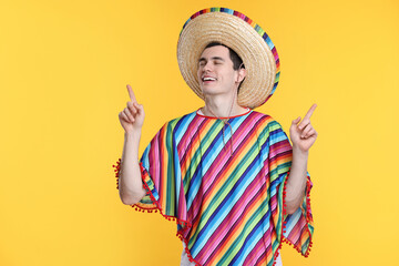 Young man in Mexican sombrero hat and poncho pointing at something on yellow background - obrazy, fototapety, plakaty