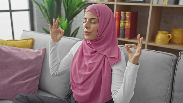 Peaceful picture of a young, attractive hispanic woman relaxing on her sofa at home, eyes closed in a healing yoga meditation pose, wearing a zen hijab