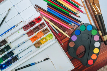 Top view of a multicolored watercolor palette, brushes, colored pencils and equipment for painting on white sheets of paper. Copy space, mockup.