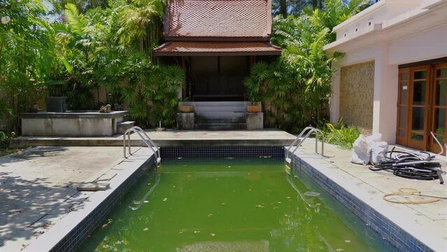 Modern tropical villa with unattended swimming pool in need of cleaning and maintenance, surrounded by lush greenery and equipped with outdoor facilities. Home care and maintenance issues.