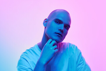 Portrait of young man with bald head, shaved face, inn casual t-shirt posing on gradient blue pink background in neon light. Concept of male beauty, body, youth, fitness, sport, health
