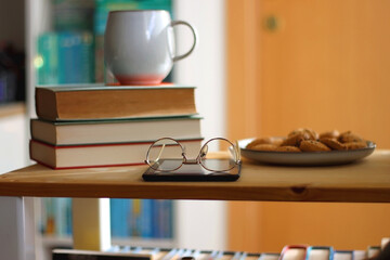 Cup of tea or coffee, pile of books, plate of cookies, reading glasses, e-reader and pen on the table. Colorful rainbow bookshelf in the background. Selective focus.