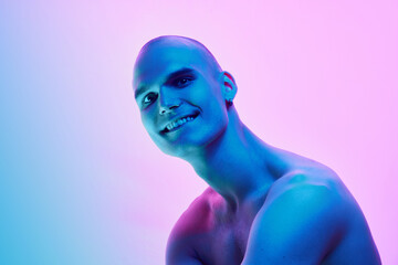 Handsome young man with bald head, shaved face posing shirtless on gradient blue pink background in neon light. Concept of male beauty, body, youth, fitness, sport, health