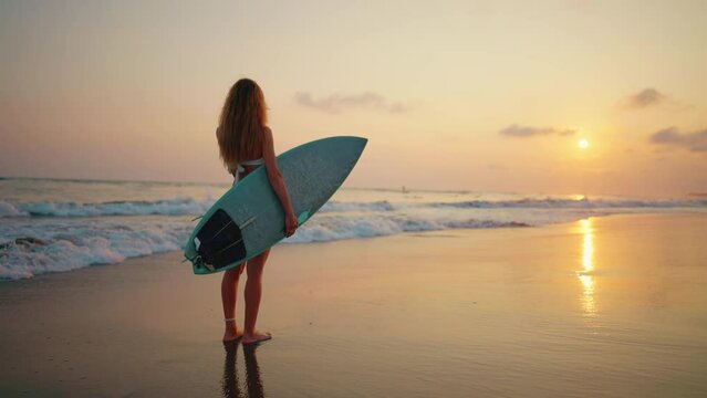Tourist woman surfer in swimsuit with surfboard stands on ocean sandy beach at sunset admires waves. Summer sport activity on tropics resort. Athlete girl. Extreme water sport surfing on vacation.