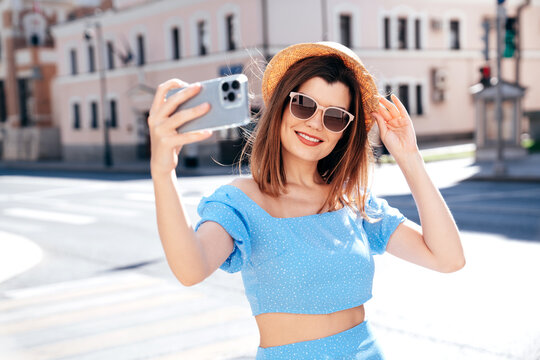Young beautiful smiling hipster woman in trendy summer blue clothes. Sexy carefree woman posing on the street background at sunset. Positive model outdoors. Cheerful and happy and hat, takes selfie