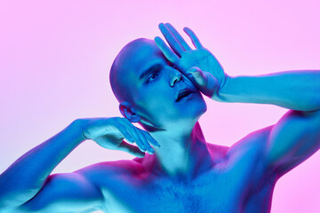 Bald man with eyes closed, hand on forehead posing on gradient blue pink background in neon....