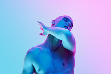 Portrait of young shirtless bald man posing with gestures on gradient blue pink background in neon...