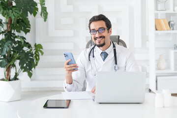 Close up portrait of smiling male doctor sitting at the desktop and talking to phone in the office...