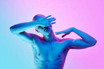 Portrait of young shirtless bald man posing with gestures on gradient blue pink background in neon light. Concept of male beauty, body, youth, fitness, sport, health
