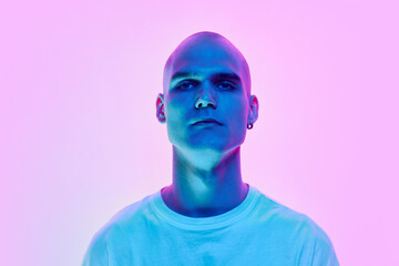 Portrait of serious young man with bald head, shaved face and ear piercing posing on gradient blue pink background in neon light. Concept of male beauty, body, youth, fitness, sport, health