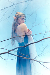 A young woman in a blue dress, resembling an elf princess, stands gracefully in front of a majestic tree.