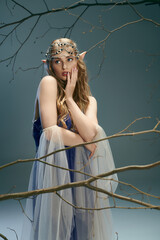 A young woman, resembling an elf princess, strikes a pose in a blue dress in a magical studio setting.