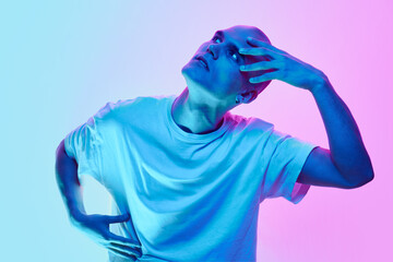 Portrait of young man with bald head posing in white casual t-shirt on gradient blue pink background in neon light. Concept of male beauty, body, youth, fitness, sport, health