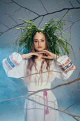 A mystical young woman dressed in white delicately balances a plant on her head in a magical studio setting.