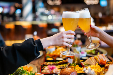 Two hands toasting with beer glasses over a table filled with assorted food in a vibrant bar atmosphere