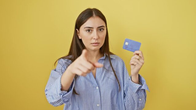 Confident young hispanic woman pointing straight at you with credit card in hand - a serious gesture on yellow isolated background.