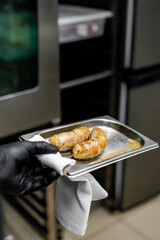 Person in black gloves holding a tray with two freshly grilled sausages, ready to serve. Background shows a blurred industrial kitchen environment.