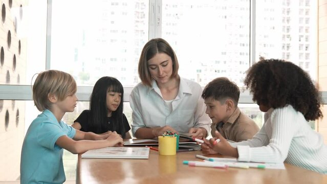 Professional caucasian teacher telling story to diverse student while sitting at table with storybook and colored book. Smart learner listening story while colored picture from instructor. Erudition.