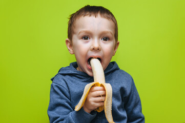 A little funny cheerful boy bites a banana, makes faces and plays around. Studio photography with green background.