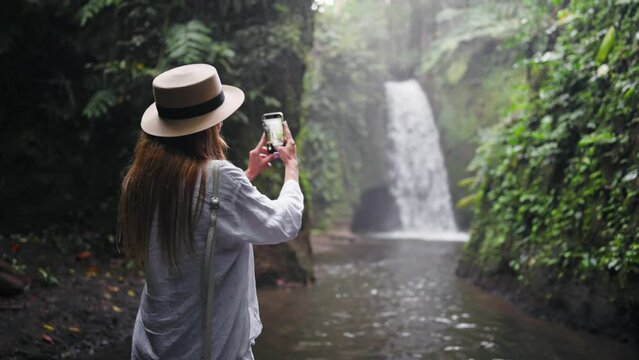 Carefree blonde woman in straw hat take photos breathtaking mountain waterfall in wild nature. Girl tourist in hike, back view. Hiking, travel, admiring tourism, vacation, peaceful journey concept.