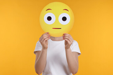 Woman covering face with surprised emoticon on yellow background